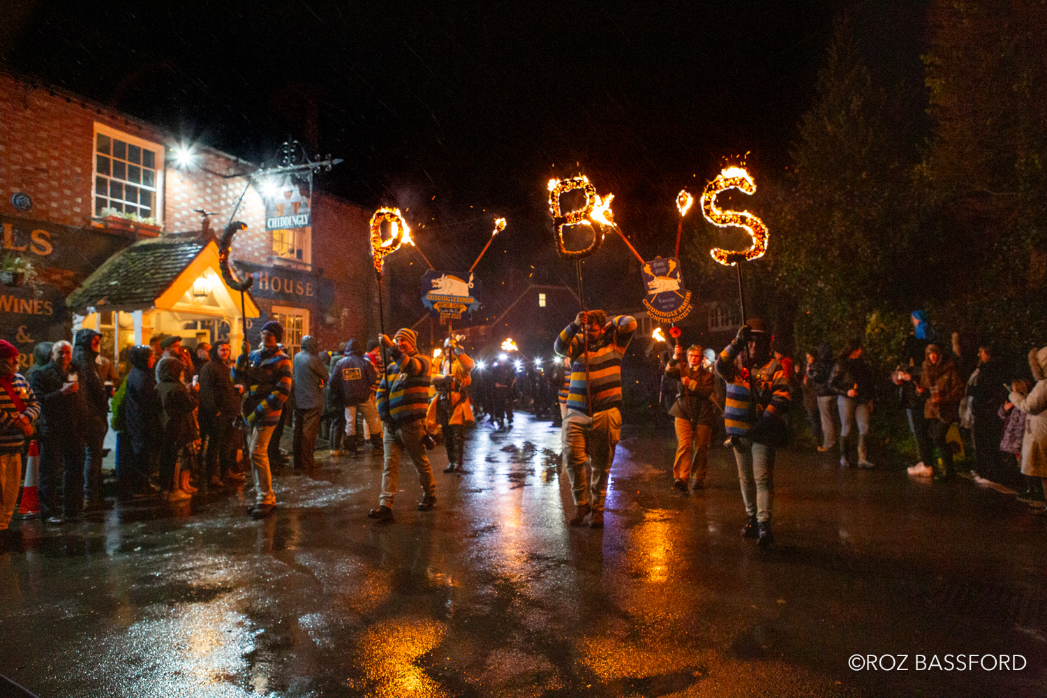 Torchlit procession at the Chiddingly Bonfire celebrations, with participants carrying illuminated bonfire society signs through village streets lined with spectators.